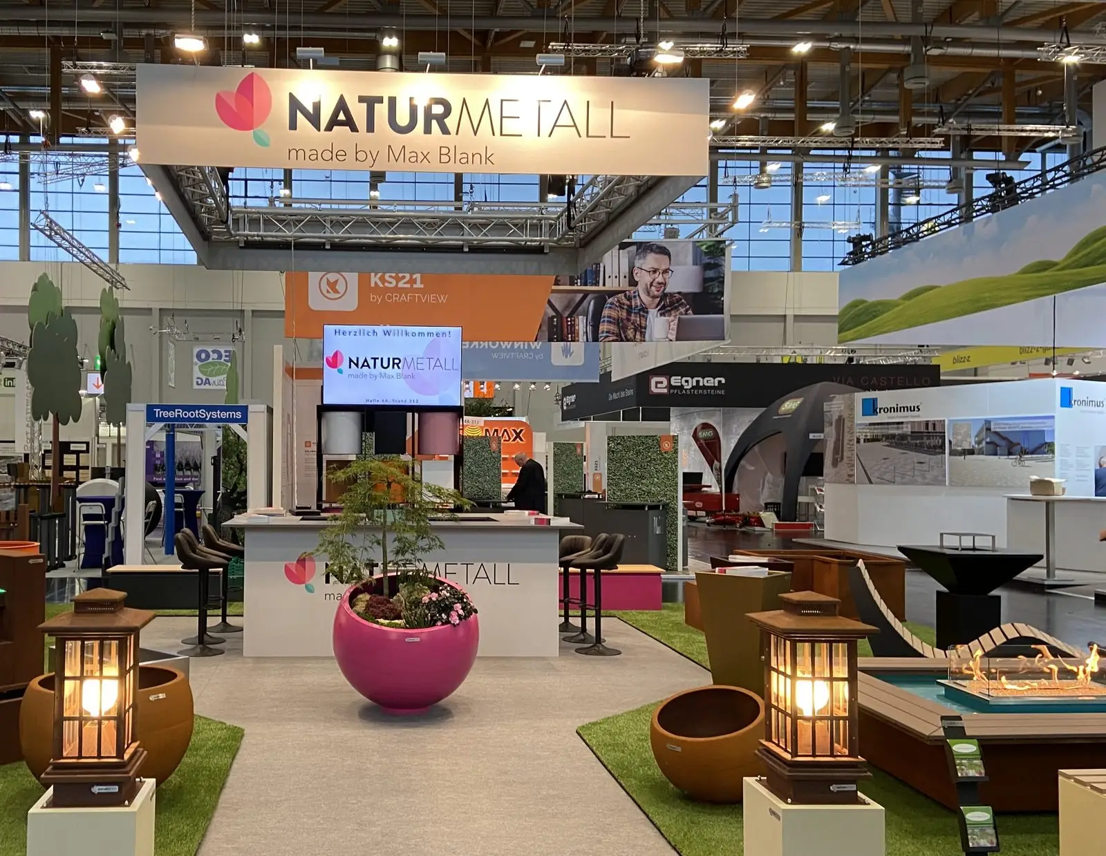 Messestand von naturmetall auf der GaLaBau 2022 mit großem Logo-Banner, Theke, Kugelpflanzgefäß in Magenta und zwei Gartenlaternen