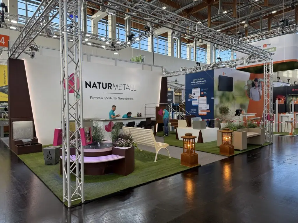 Messestand von naturmetall auf der GaLaBau 2026 mit großer Logowand, Theke, rundem Pflanztrog mit Sitzbank und Quellbrunnen, Gartenbank und Gartenlaternen